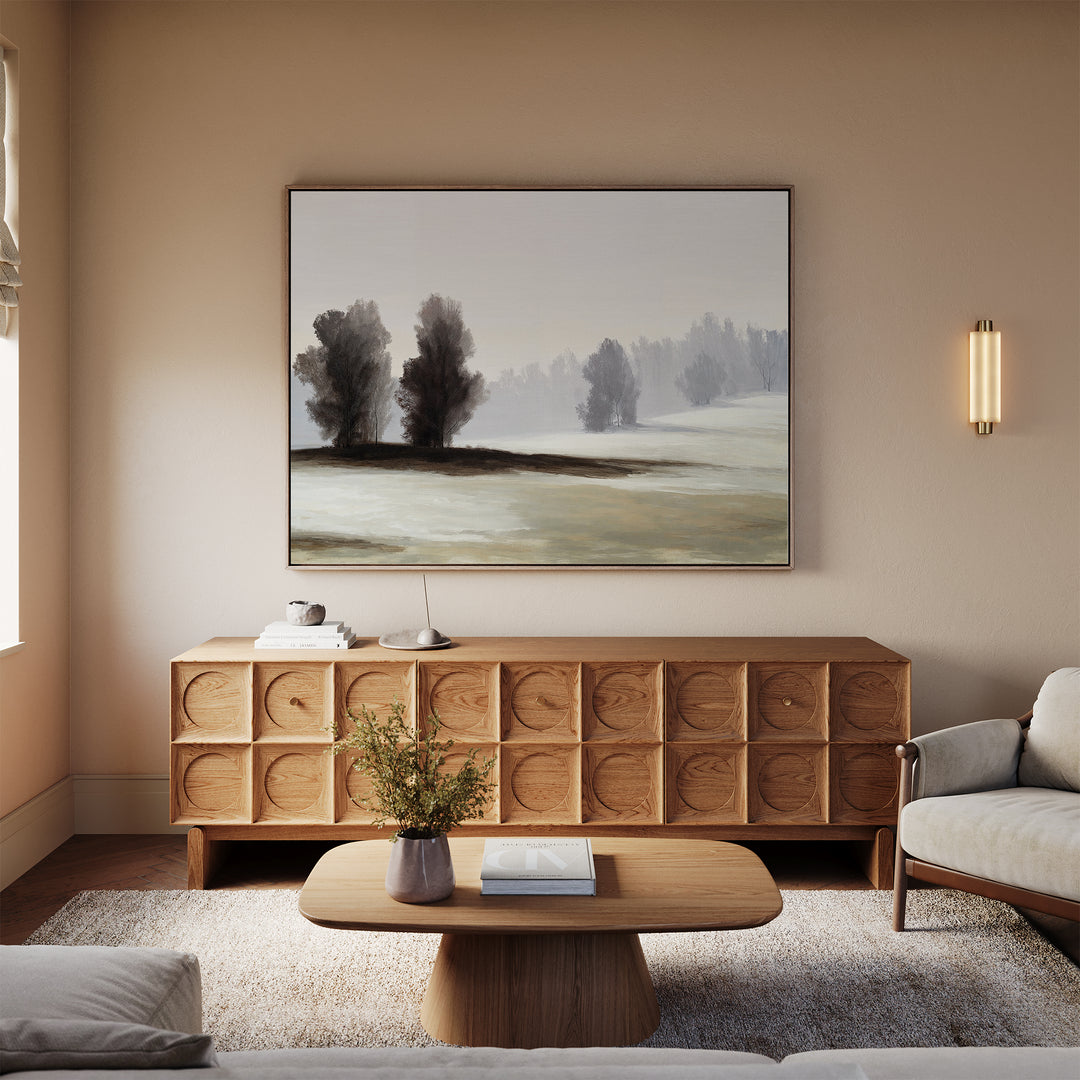 A cozy living room with neutral tones features a wooden credenza, beige sofa, and light wood coffee table. The Winter Dusk Canvas Art and minimalist wall decor complement a modern wall sconce and a white rug.