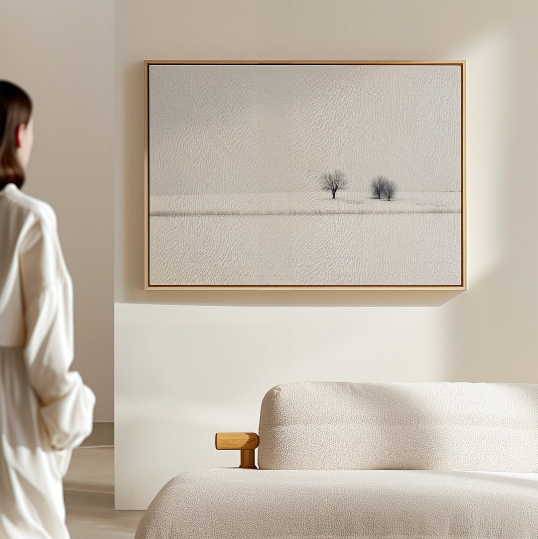 A person stands in a light-filled, minimal room with a beige sofa and winter decor, featuring the Winter Silence Canvas Art—a minimalist wall piece showing a serene, snow-covered landscape with two distant trees.