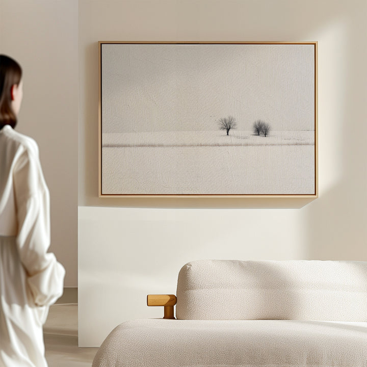 A person stands in a light-filled, minimal room with a beige sofa and winter decor, featuring the Winter Silence Canvas Art—a minimalist wall piece showing a serene, snow-covered landscape with two distant trees.