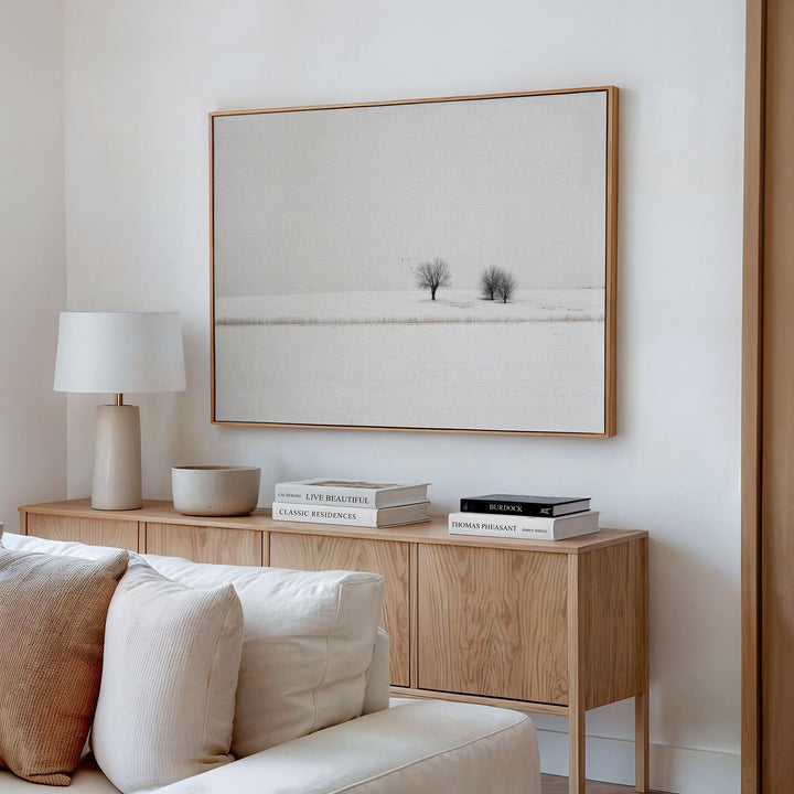 A minimalist living room features a light wood sideboard, white table lamp, stacked books, and a bowl. Above hangs the Winter Silence Canvas Art depicting a snowy landscape with two bare trees. Neutral tones complete the decor.