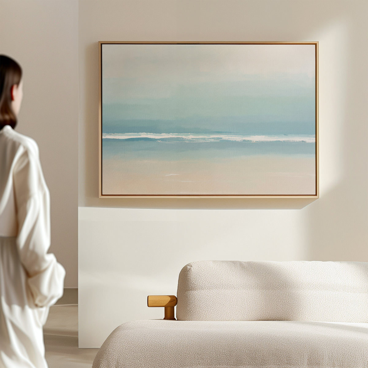 A person in white stands by a beige sofa, admiring the Airy Shore Canvas Art - Horizontal depicting a beach and ocean on a light wall in a serene, modern living room.