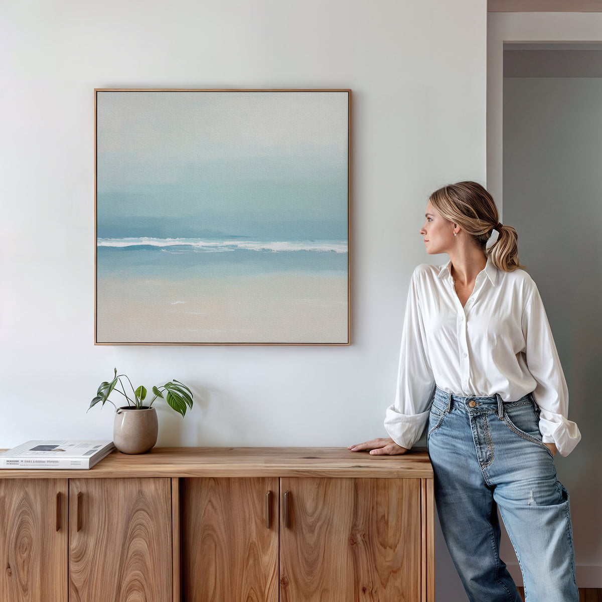 A woman in a white blouse and jeans leans on a wooden cabinet, admiring the Airy Shore Canvas Art - Square, a framed seascape on the white wall. A small plant and books add to the cozy coastal decor atop the cabinet.