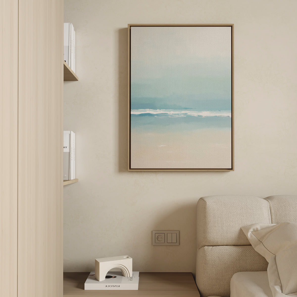 A minimalist living space with a beige sofa, light wooden shelves holding books, and the Airy Shore Canvas Art - Vertical coastal sea print in calming blue, white, and tan tones hanging above an electrical outlet.