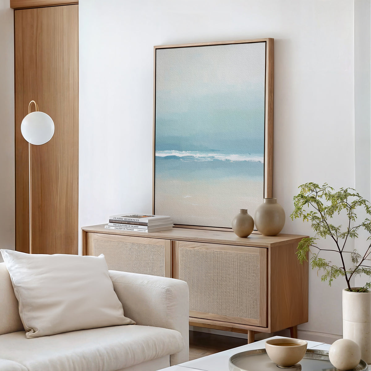 A minimalist living room with a light wood sideboard, ceramic vases, books, a white sofa with pillows, and a round floor lamp. Airy Shore Canvas Art - Vertical brings a seaside vibe above the small potted plant on the white table.