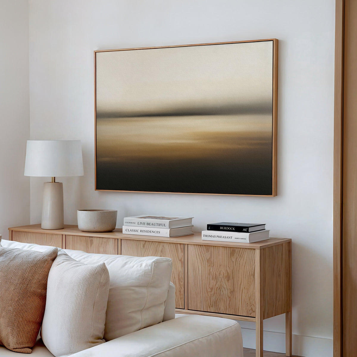 A modern living room with a beige sofa, wooden sideboard, white lamp, stacked books, and the Amber Fields Canvas Art - Horizontal featuring neutral and amber tones hanging on the wall.