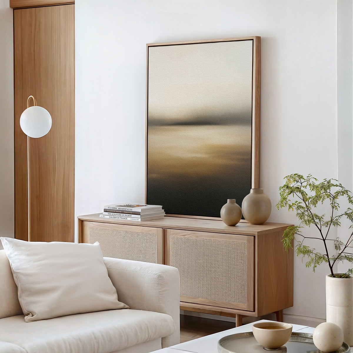 A minimalist living room featuring the Amber Fields Canvas Art - Vertical, a light wood sideboard, neutral vases, stacked books, a white sofa with cushions, amber accents, a small plant, and a round white lamp by the wooden door.