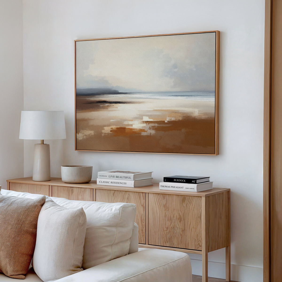 A modern living room features a light wood sideboard with books, lamp, and bowl. Above it hangs the Amber Shore Canvas Art - Horizontal, a large abstract landscape in soft beige and blue. A cream sofa sits in the foreground.