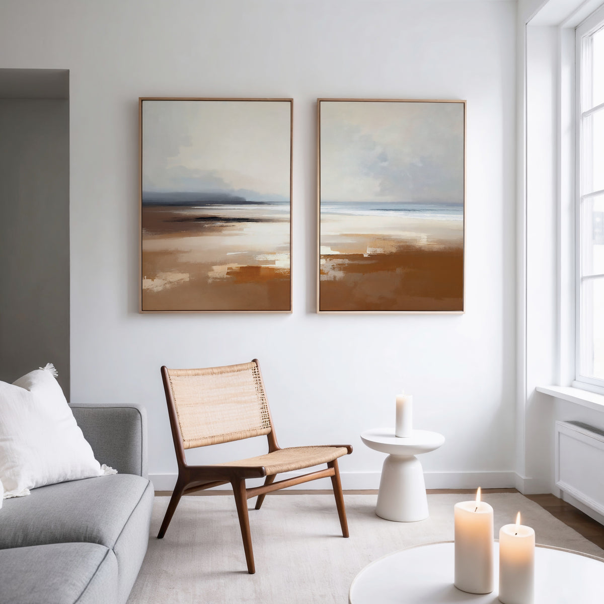 A modern, minimal living room features a light gray sofa, wooden chair, and the "Amber Shore - Set Of 2" abstract landscape paintings. A small white side table and lit candles add coziness as natural light streams through large windows.