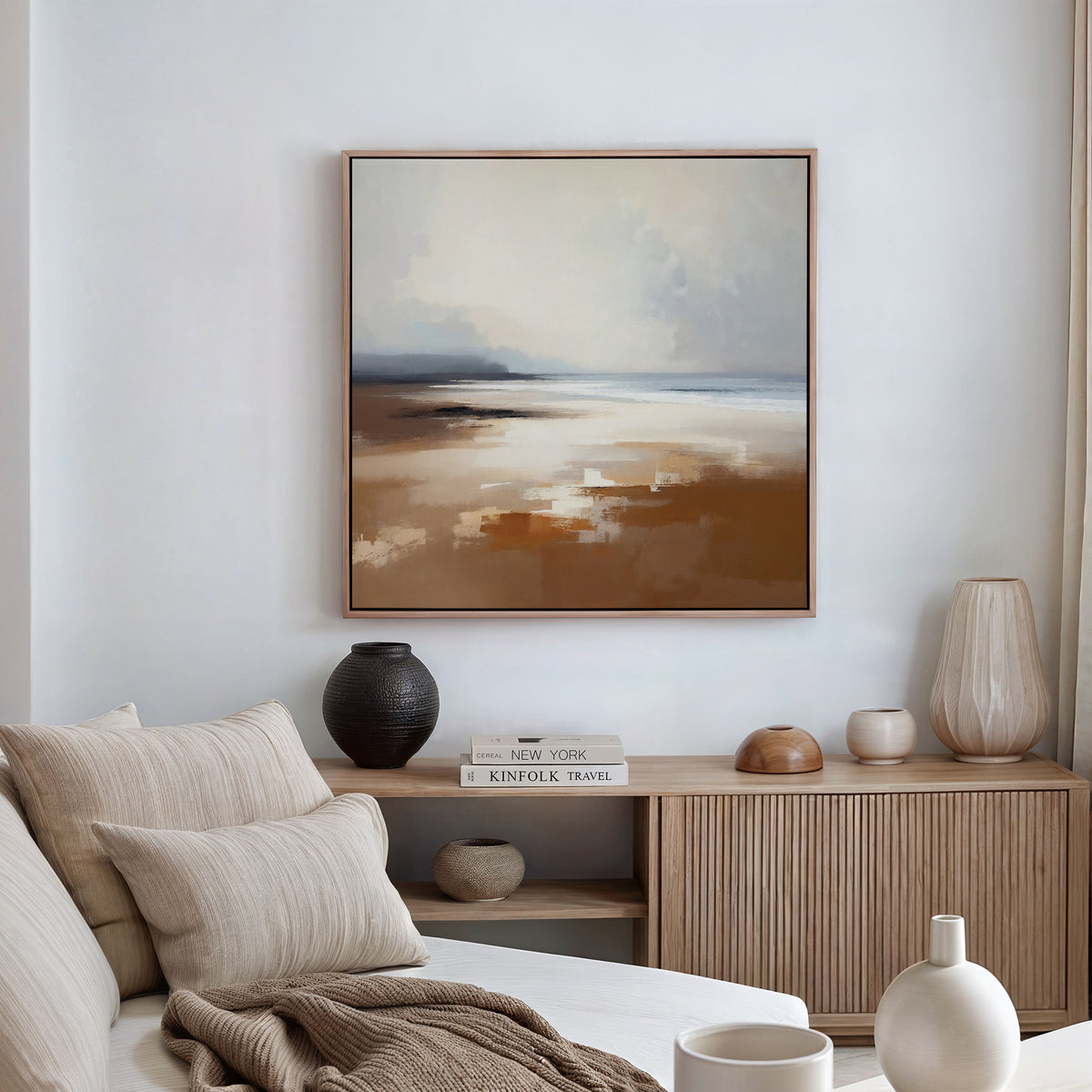 A modern living room in neutral tones features a beige sofa with cushions, a wooden sideboard with vases and books, and the Amber Shore Canvas Art - Square—an abstract landscape—displayed above as tranquil wall decor.