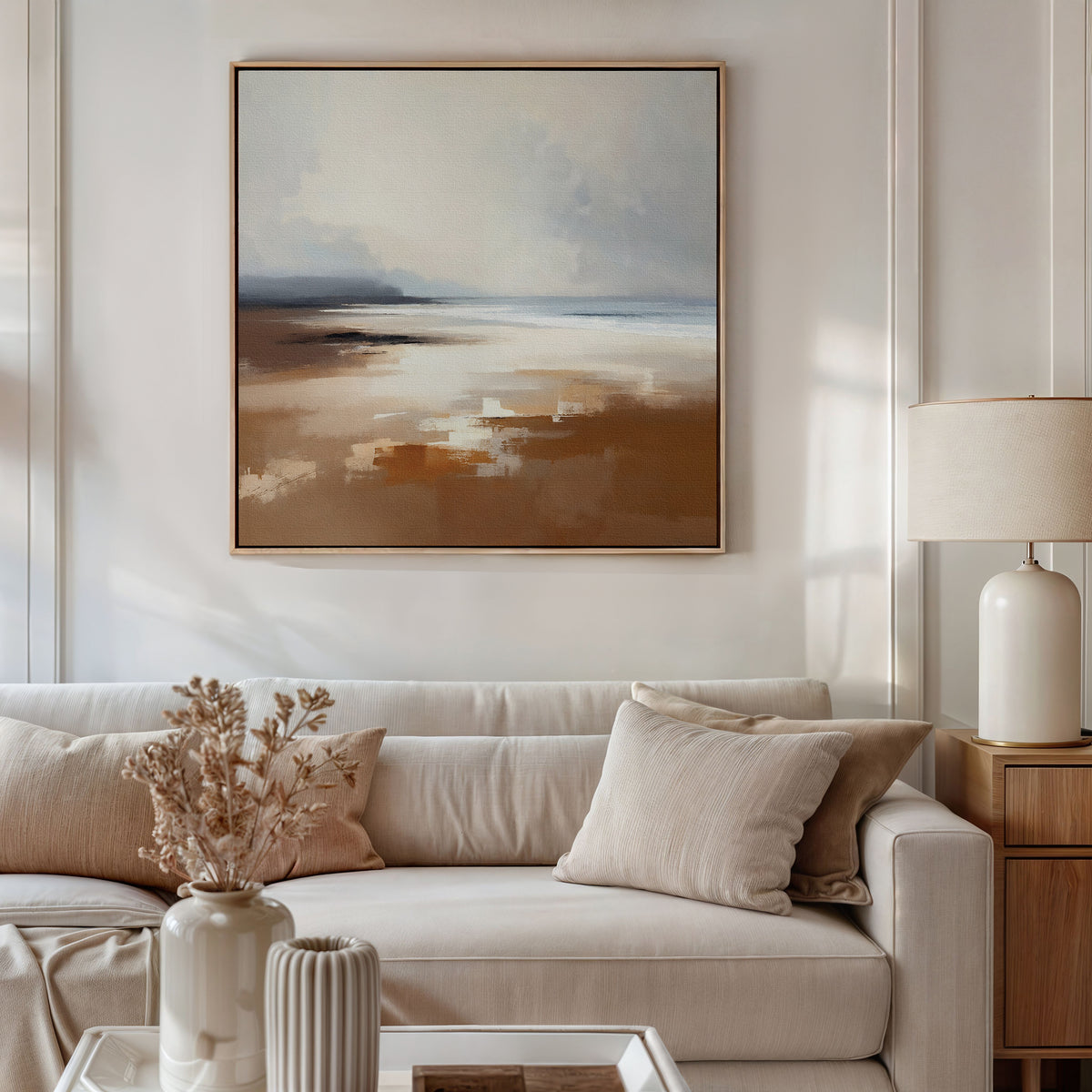 A modern living room featuring a beige sofa, neutral cushions, dried flowers in a vase, a table lamp, and the Amber Shore Canvas Art - Square with soft brown, beige, and blue tones for serene elegance.