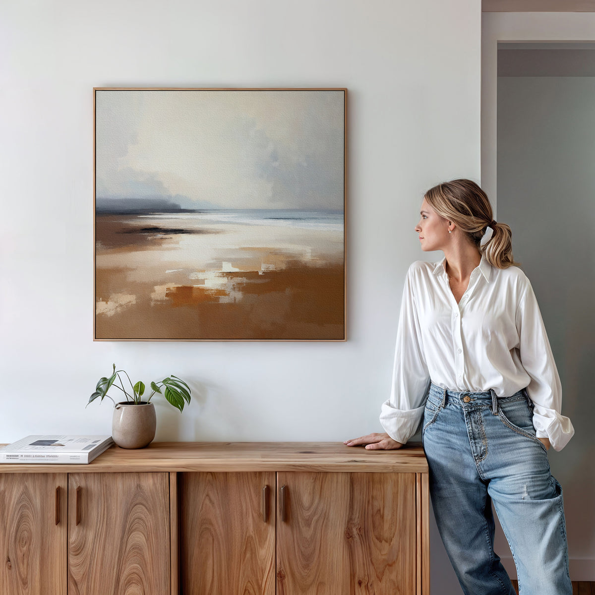 A woman in a white blouse and blue jeans stands by a wooden cabinet, gazing at the Amber Shore Canvas Art - Square. A plant and a book rest on the cabinet, enhancing the calm, Nordic Park-inspired atmosphere.