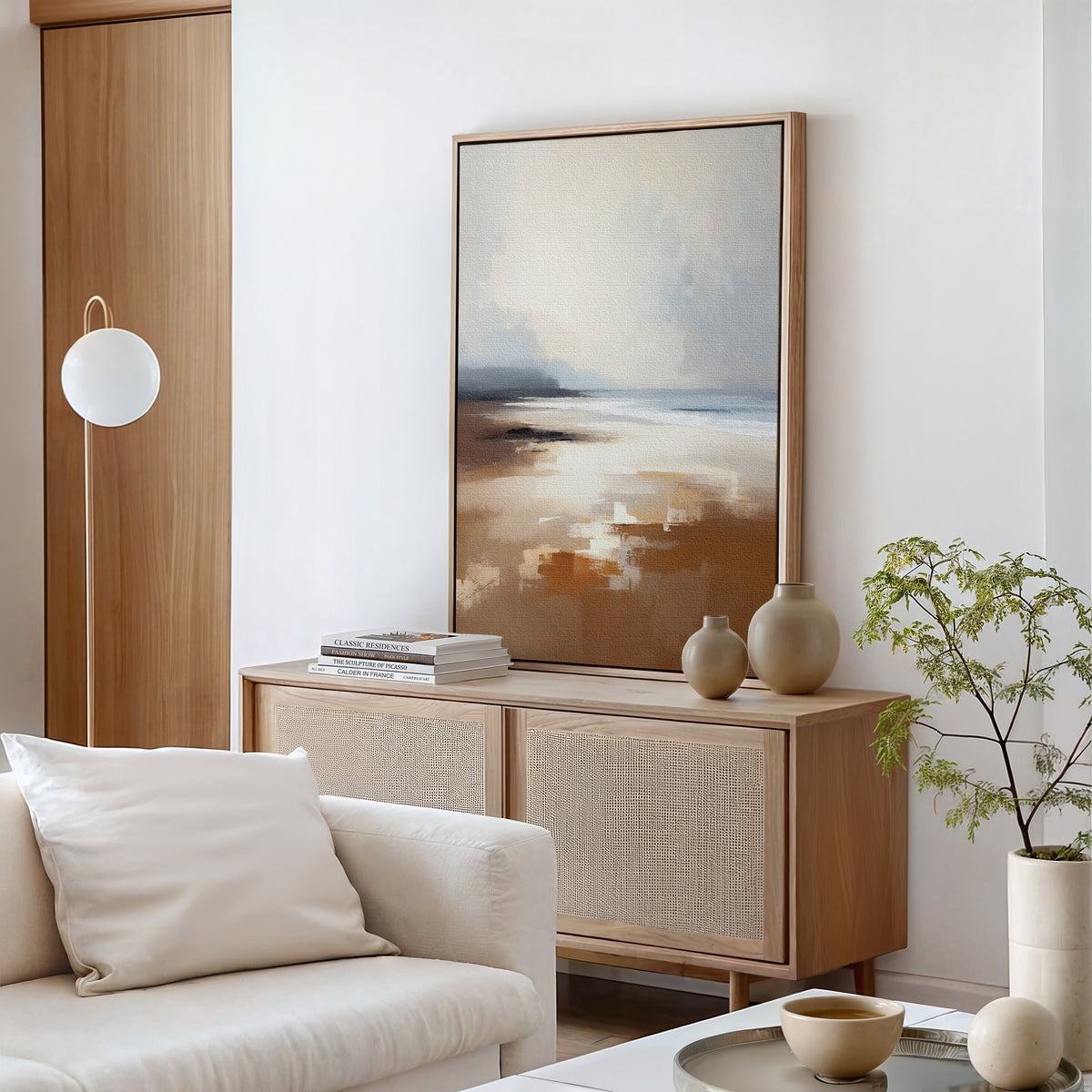 A modern living room featuring the Amber Shore Canvas Art - Vertical, a light wood sideboard, stacked books, ceramic vases, a small potted plant, a round floor lamp, and a white sofa with neutral cushions.