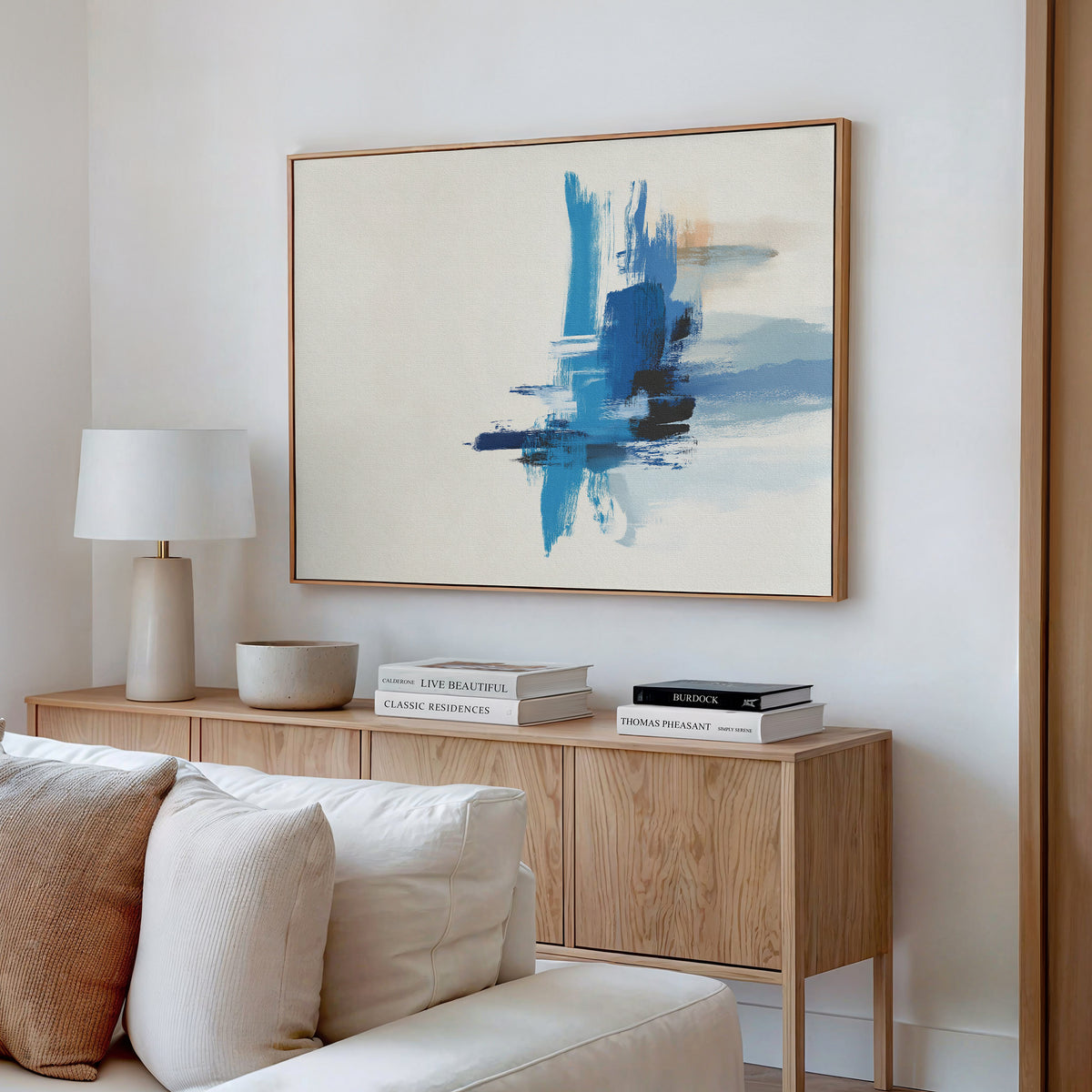 A modern living room features a light wooden sideboard, a white table lamp, stacked books, and the Azure Convergence Canvas Art - Horizontal in blue tones displayed above a white sofa with cream cushions.