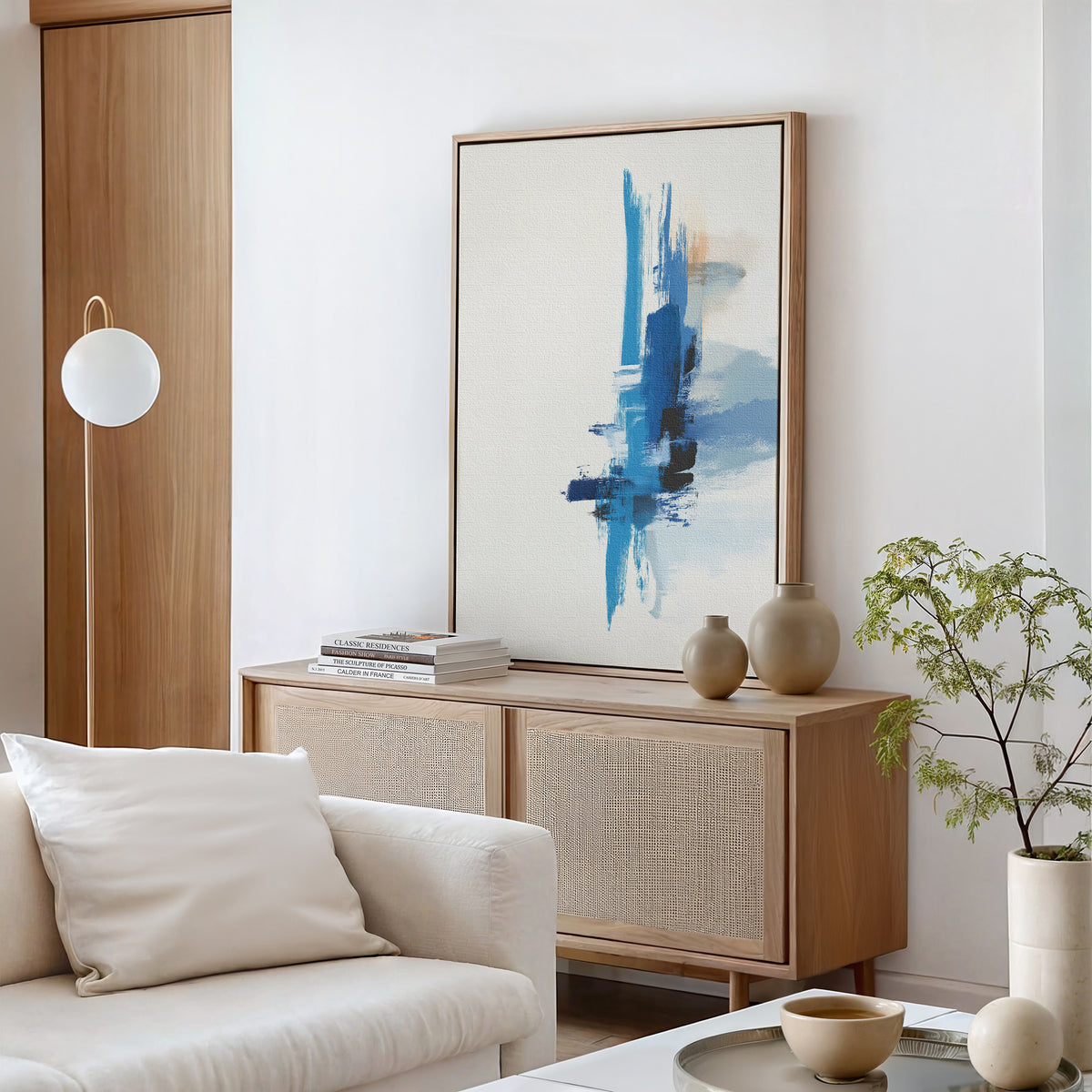 Minimalist living room with light wood furniture, the Azure Convergence Canvas Art - Vertical in blue and white, neutral vases, stacked books, a potted plant, and a white sofa accented with cream cushions.