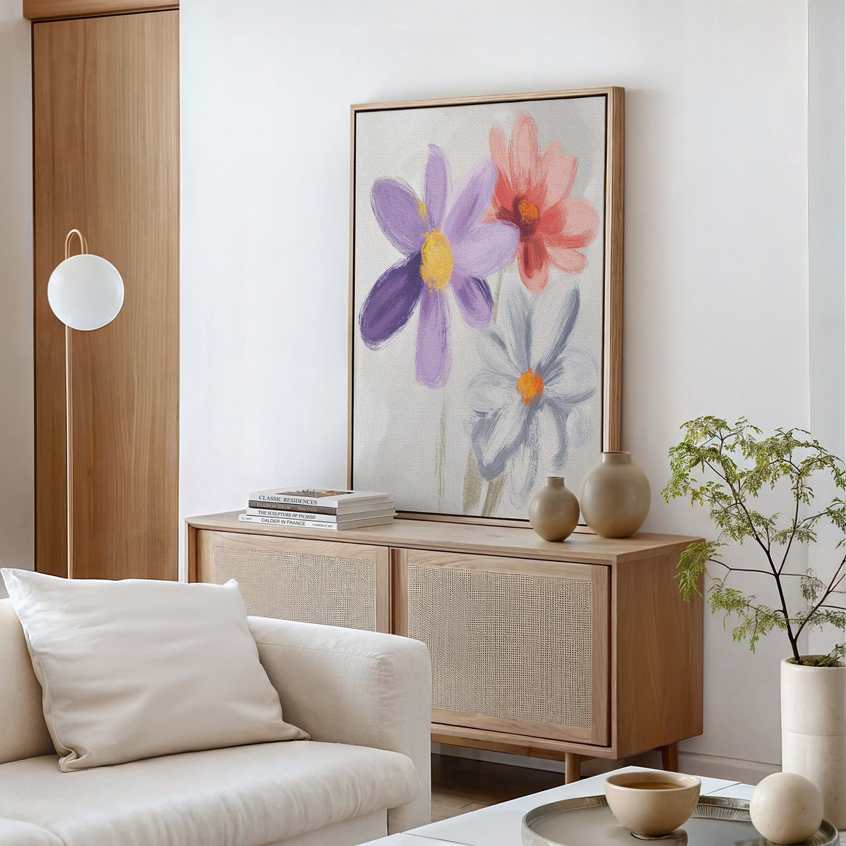 A modern living room features a light wooden sideboard, Blooming Harmony Canvas Art - Vertical with three colorful flowers, neutral-toned vases, books, a white sofa with pillows, and a small green plant on the coffee table.