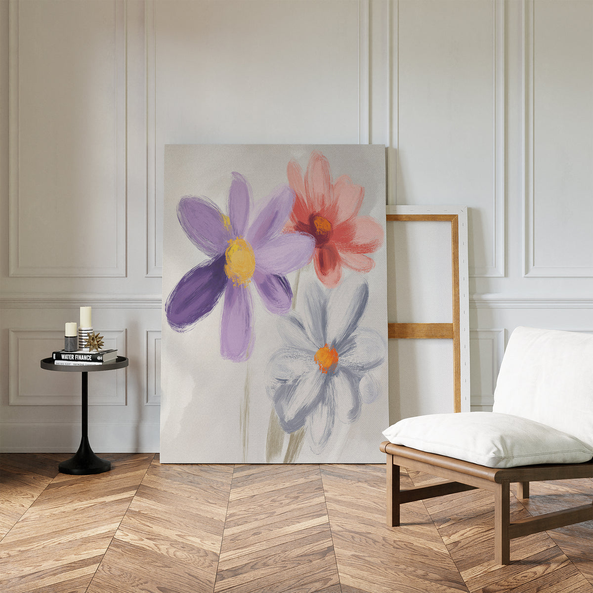 The Blooming Harmony Canvas Art - Vertical, featuring three painted flowers, leans against a white paneled wall in a stylish room with herringbone wood floors, a modern low chair, and a small black round table with candles and books.