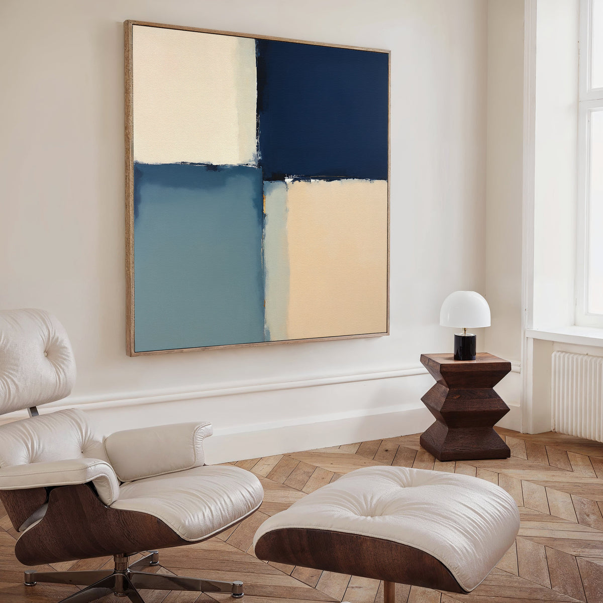 A modern living room with a white leather lounge chair and ottoman, a geometric wooden side table and lamp, and the Blue Quarters Canvas Art - Square in blue, cream, and beige tones displayed on the wall.
