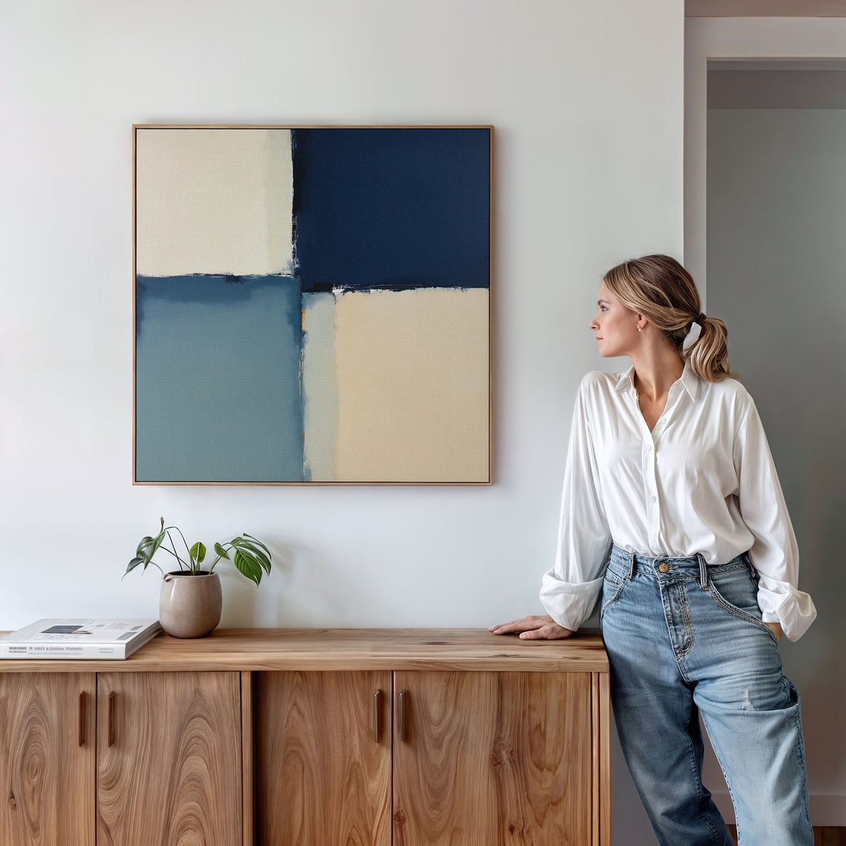 A woman in a white blouse and jeans leans against a wooden cabinet, gazing at the Blue Quarters Canvas Art - Square, featuring blue and beige shapes. A plant and books on the cabinet create a modern living room vibe.