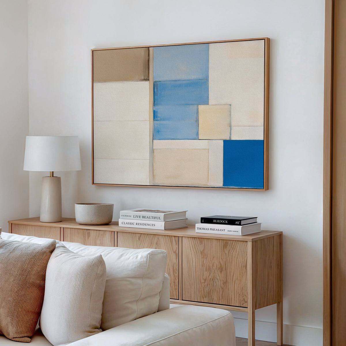 A modern living room features a wooden sideboard with books, a lamp, and a decor bowl. Above it hangs the Blue Tiles Composition Canvas Art - Horizontal in beige, blue, and white. A light sofa completes the contemporary decor.