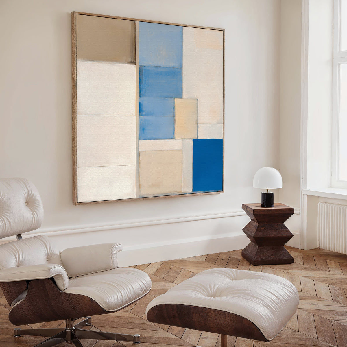 A modern living room with a white lounge chair and ottoman, a small dark wood side table with a white lamp, and the Blue Tiles Composition Canvas Art - Square featuring abstract blue, beige, and white rectangles on the wall.