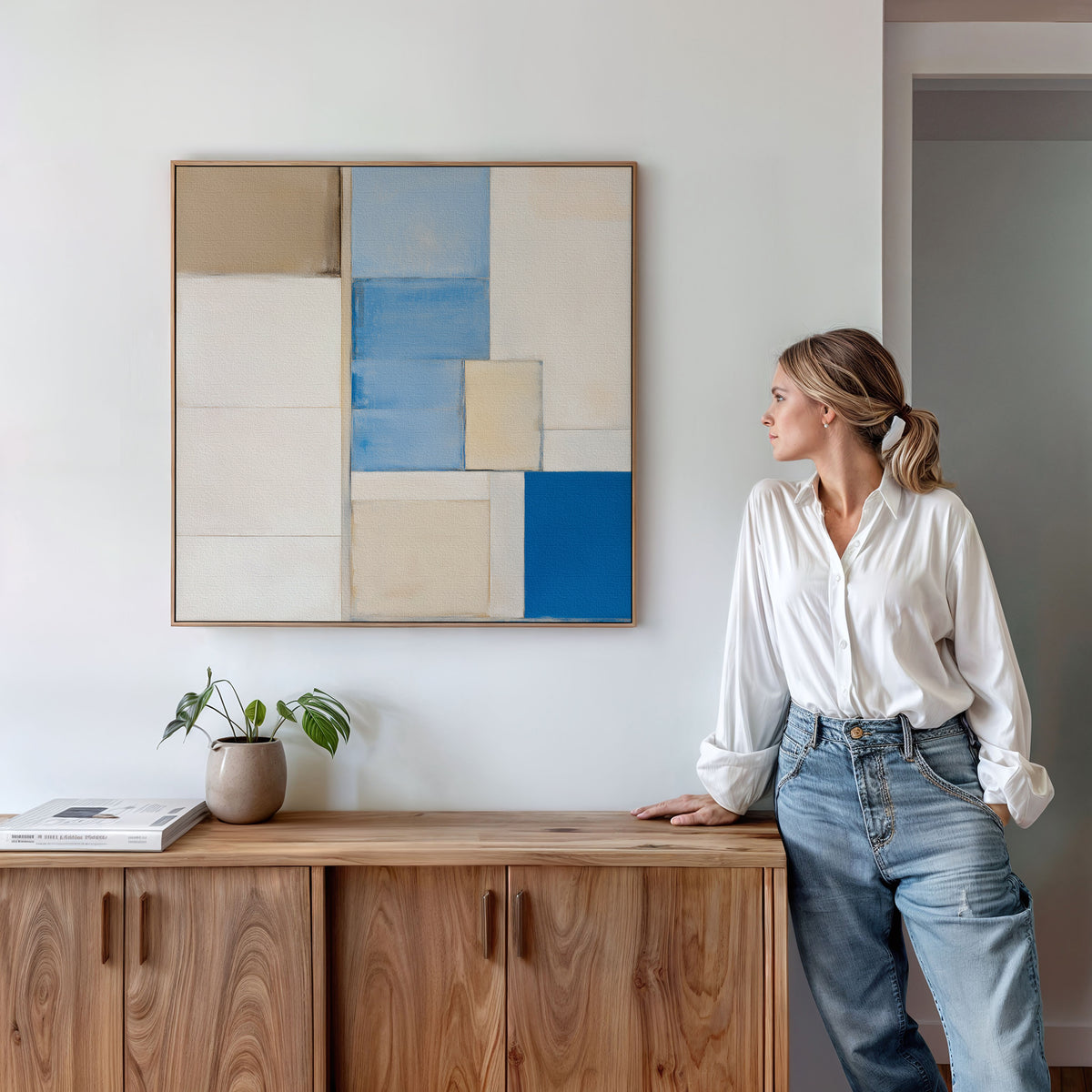 A woman in a white shirt and jeans leans against a wooden cabinet, admiring the Blue Tiles Composition Canvas Art - Square, featuring blue, beige, and white rectangles. Books and a small plant accent the modern living room decor.