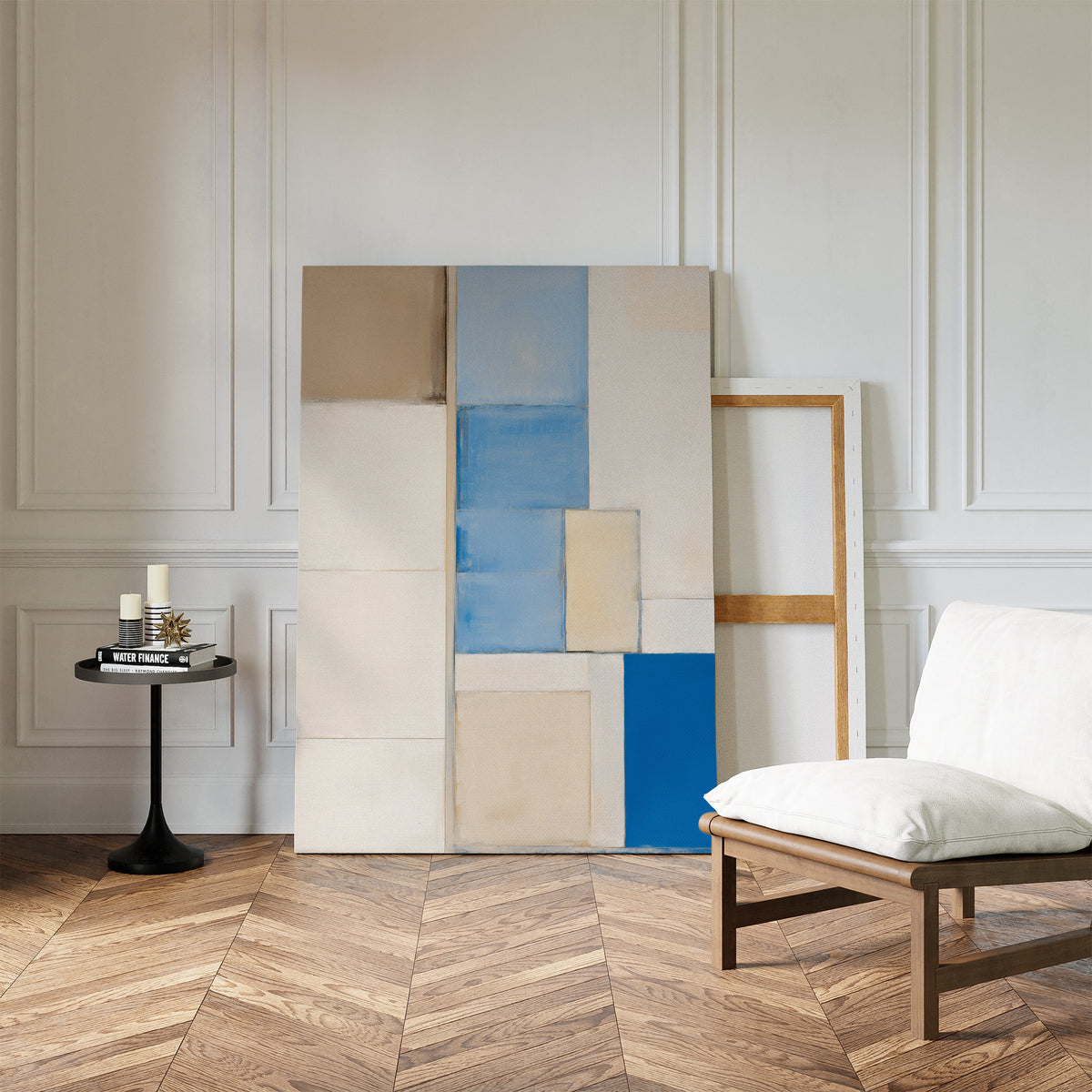 A modern space with wood herringbone floors, a white cushioned chair, side table with candles and books, and the Blue Tiles Composition Canvas Art - Vertical featuring blue, beige, and cream blocks against a crisp white wall.