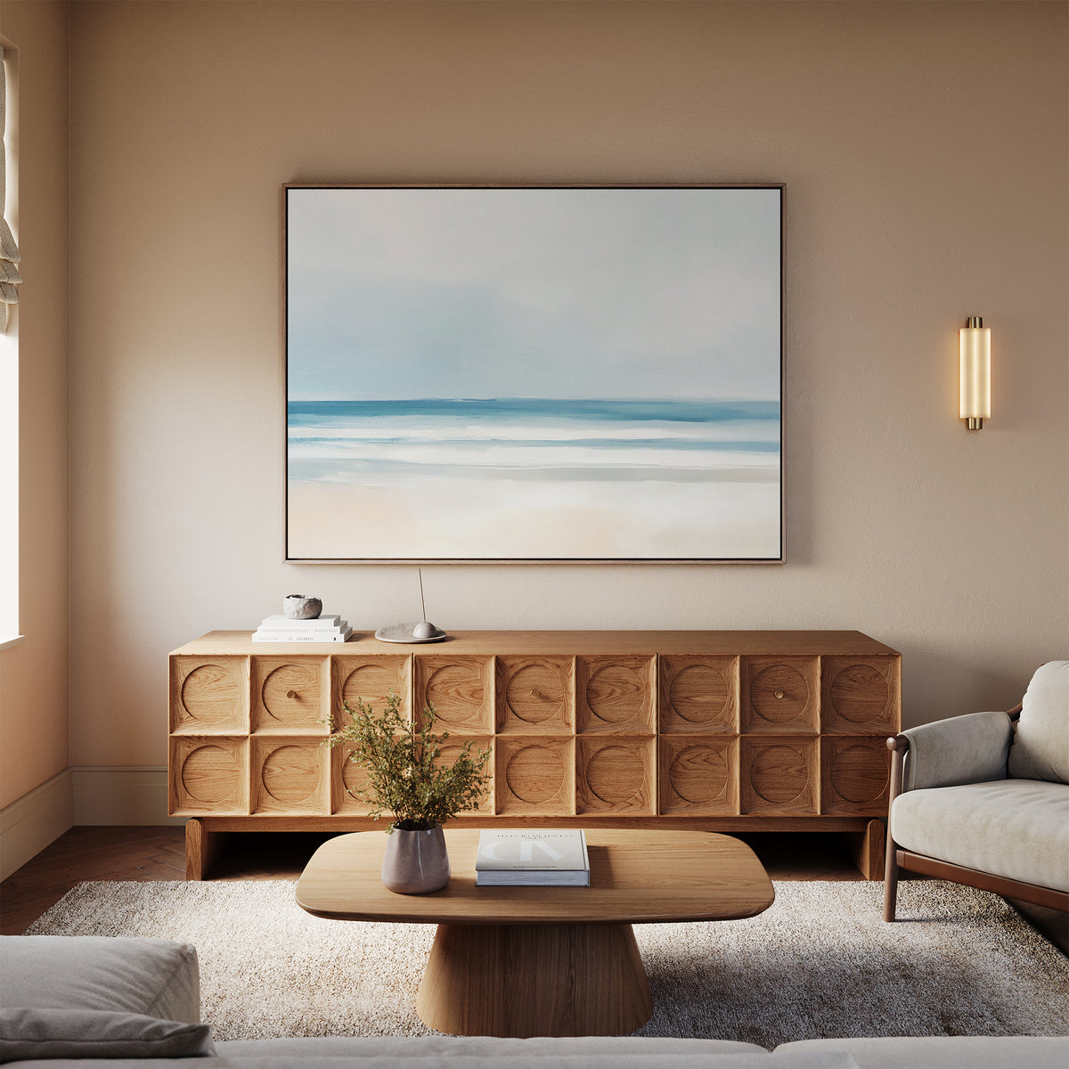 A minimalist living room featuring a light wood coffee table, textured sideboard, beige sofa, and tranquil Blurred Beach Scene Canvas Art - Horizontal. A modern sconce illuminates the wall, and a flower vase decorates the coffee table.