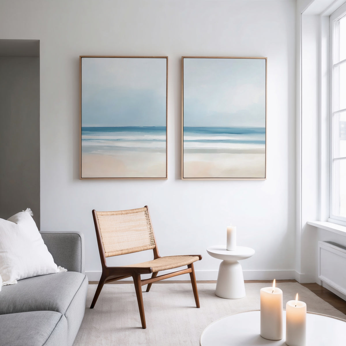 A minimalist living room features a gray sofa, woven chair, small round table, lit candles, and serene decor highlighted by the Blurred Beach Scene - Set Of 2 wall art on a white wall near a large window.