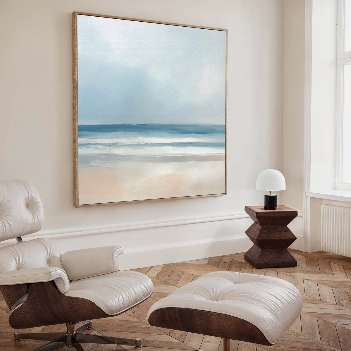 A modern, minimalist room featuring a white leather lounge chair and ottoman, a wooden side table with a white lamp, and tranquil decor—a Blurred Beach Scene Canvas Art - Square displaying a serene coastal view in soft blue and beige tones.