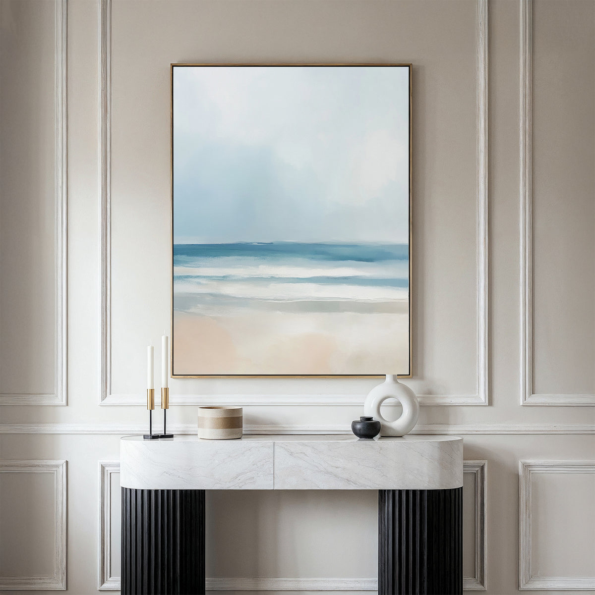 A minimalist console table with decor stands against a paneled wall, topped by the Blurred Beach Scene Canvas Art - Vertical, an abstract coastal print in soft blue, white, and beige—ideal for serene beach-inspired decor.