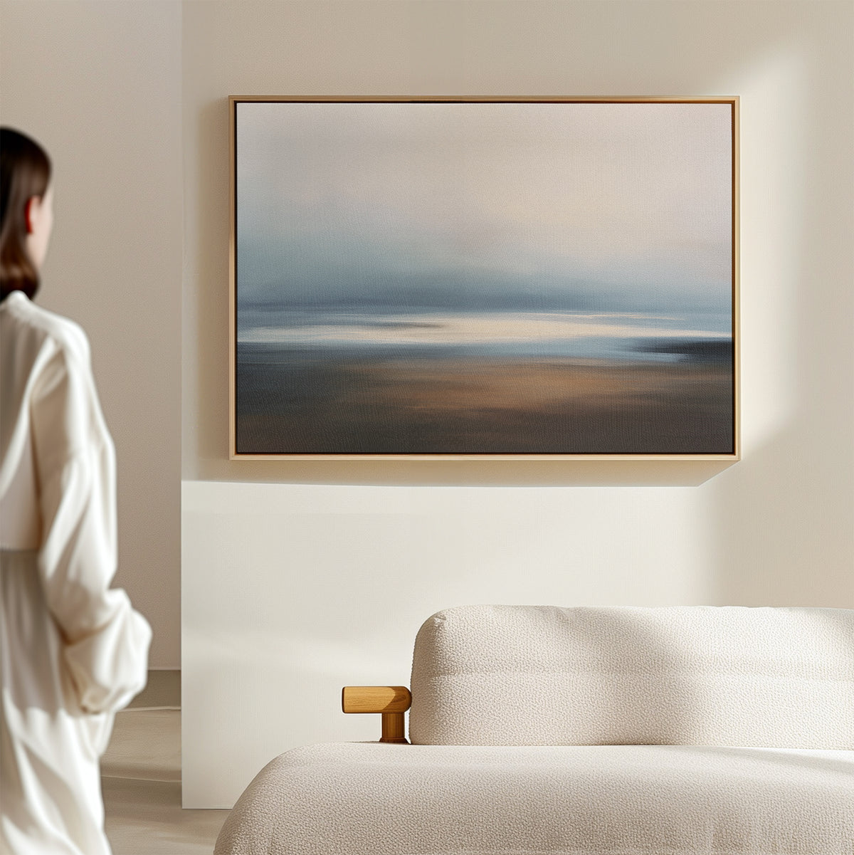 A person in a white shirt stands by a beige sofa, admiring the Blurred Seascape Canvas Art - Horizontal with soft blue, gray, and brown tones on a light wall in a minimal, modern room.