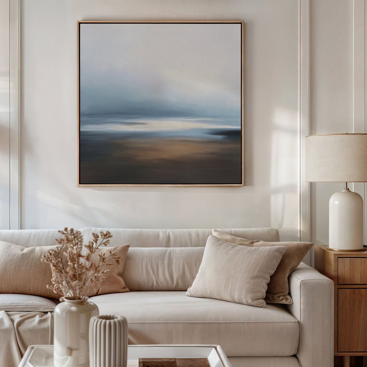 A modern living room features a beige sofa with neutral cushions, a wooden side table with a lamp, and a vase of dried flowers. The Blurred Seascape Canvas Art - Square in soft blue and brown tones decorates the wall.