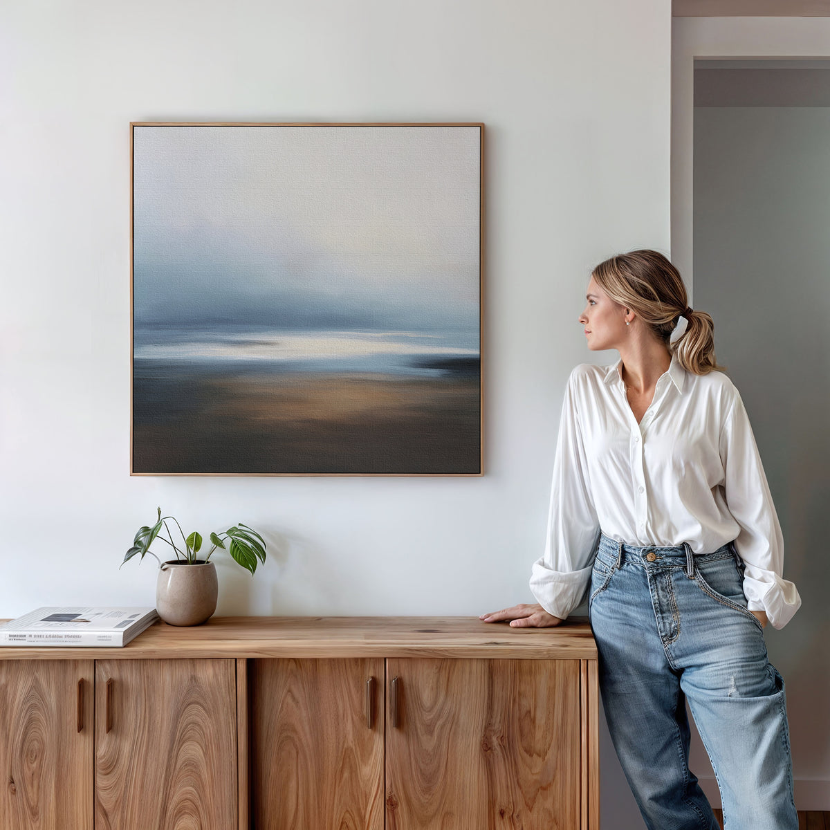 A woman in a white blouse and jeans leans on a wooden cabinet, gazing at the Blurred Seascape Canvas Art - Square above. A potted plant and books rest on the cabinet, adding to the minimalist vibe of the room.