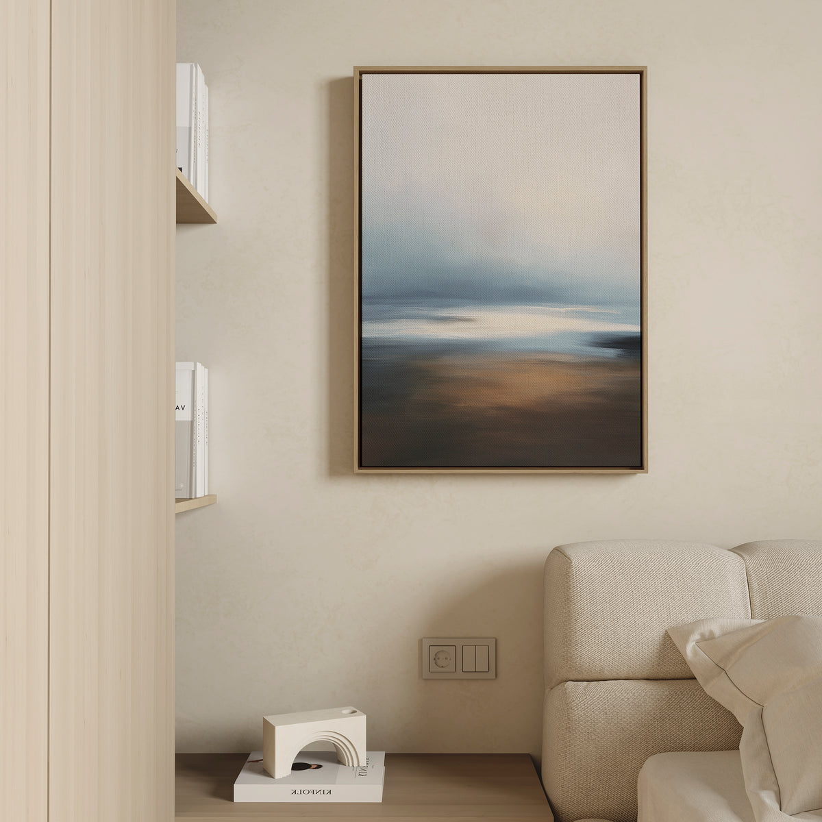 A minimalist living room corner features a beige sofa, a book shelf, a small white geometric decor piece, and the Blurred Seascape Canvas Art - Vertical on the wall for a touch of coastal serenity.