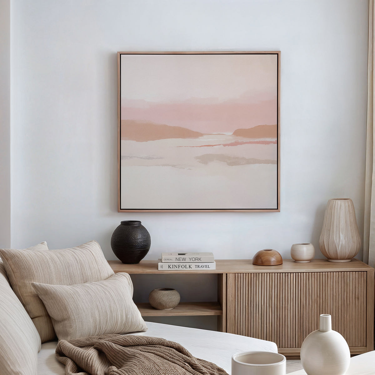 A minimalist living room features a beige sofa with neutral cushions, a wooden sideboard with vases and books, and the Blush Mesa Canvas Art - Square in calming earth tones displayed on a white wall.