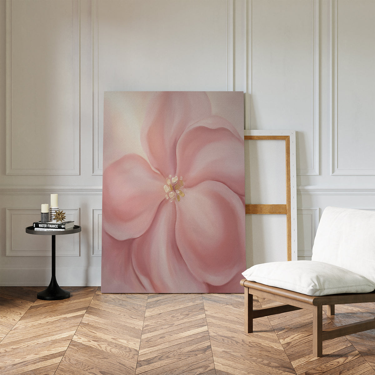 The Blushing Magnolia Canvas Art - Vertical, with its soft pink flower, leans against a white paneled wall next to an empty frame. A cushioned chair and small black table with candles and books sit nearby on a wooden herringbone floor.