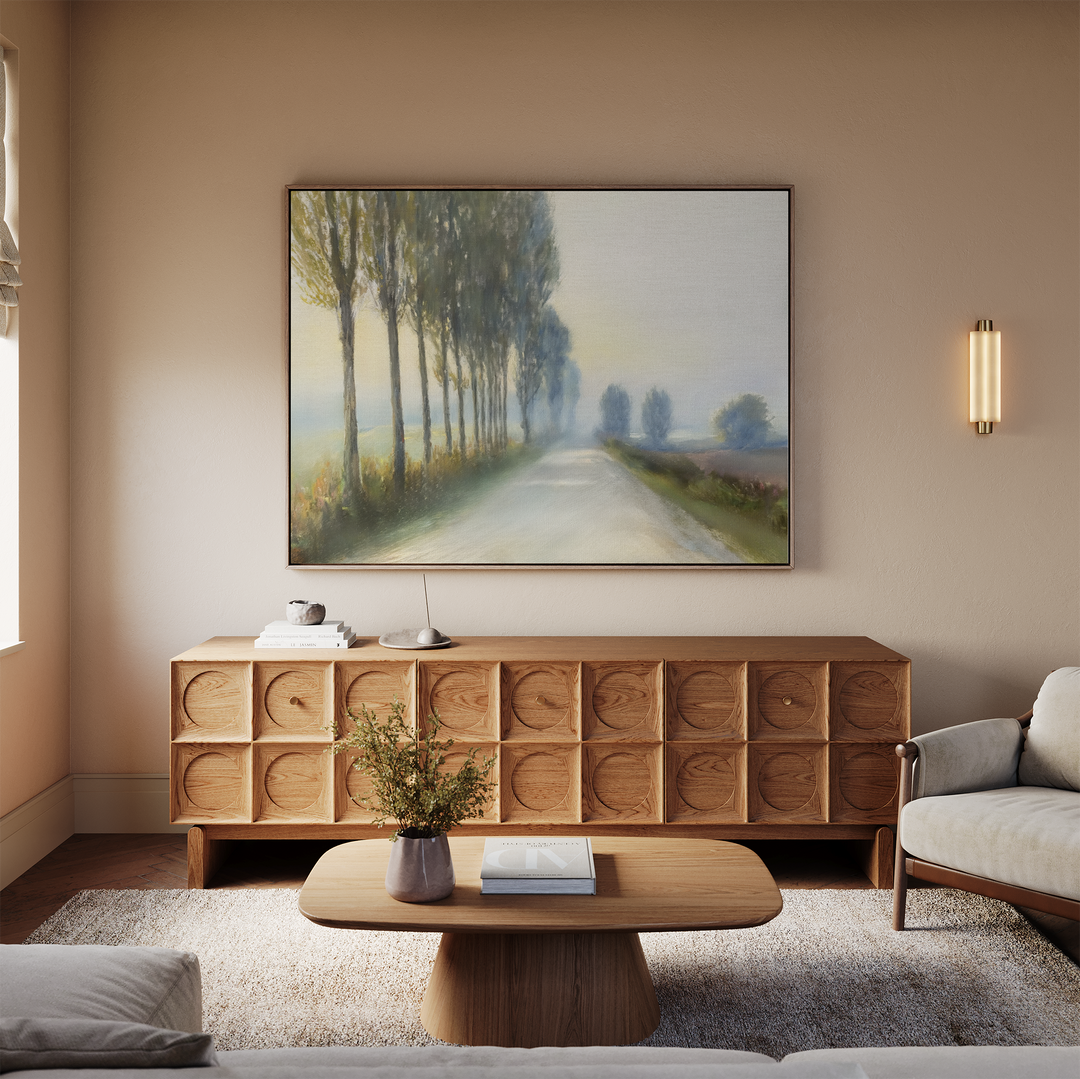A cozy living room with a wooden sideboard, coffee table, and light rug. The Forest Haze Canvas Art—a tranquil landscape—hangs above the sideboard, which is adorned with a small lamp and vase.