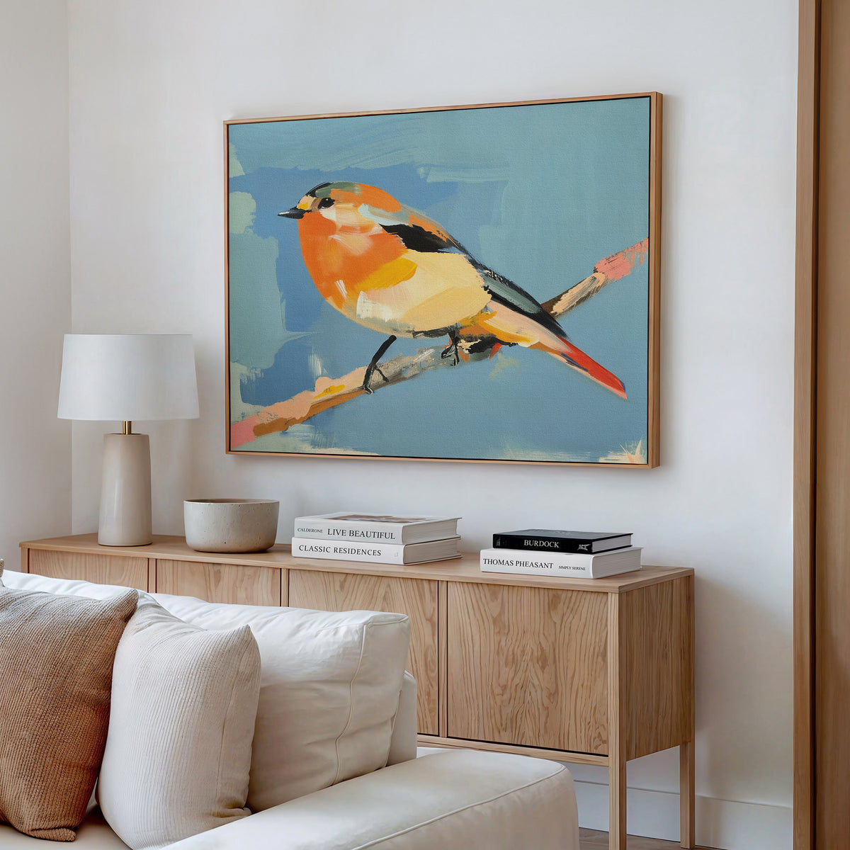 A modern living room featuring a wooden sideboard, books, lamp, bowl, and the Bright Songbird Canvas Art - Horizontal—a large colorful bird on a branch against blue—adds charm to the space.