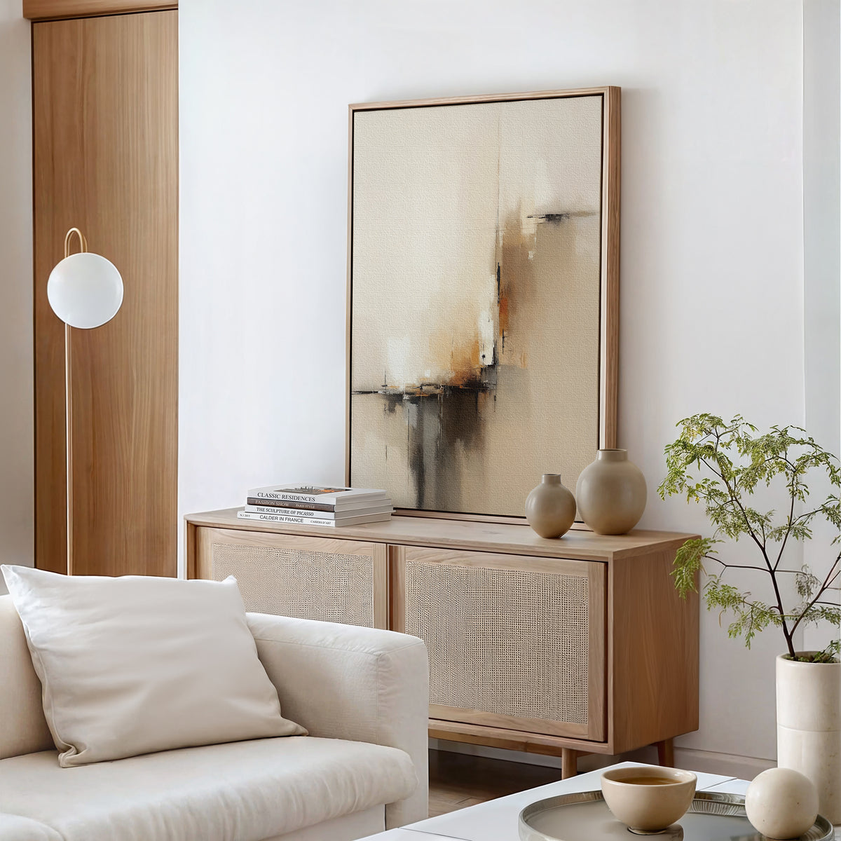 A modern, minimalist living room features a light wood sideboard, ceramic vases, stacked books, a white sofa, potted plant, round floor lamp, and the Burnt Sienna Convergence Canvas Art - Vertical for contemporary abstract-inspired elegance.
