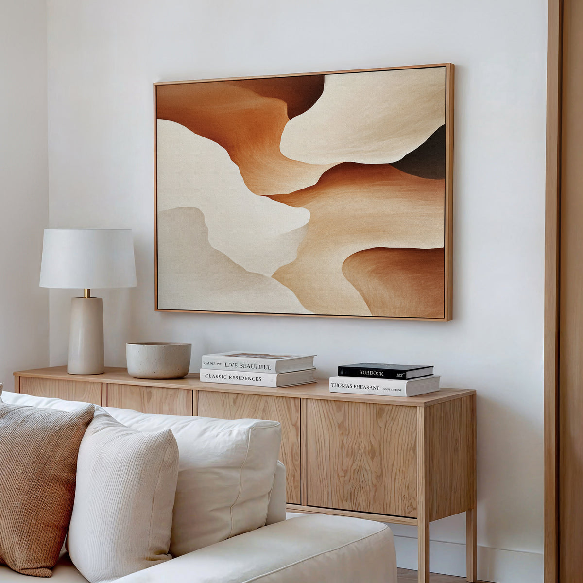 A modern, minimal living room features a wooden sideboard, neutral decor, a white lamp, stacked books, a bowl, and the Canyon Curves Canvas Art - Horizontal in warm beige, brown, and cream tones displayed above.