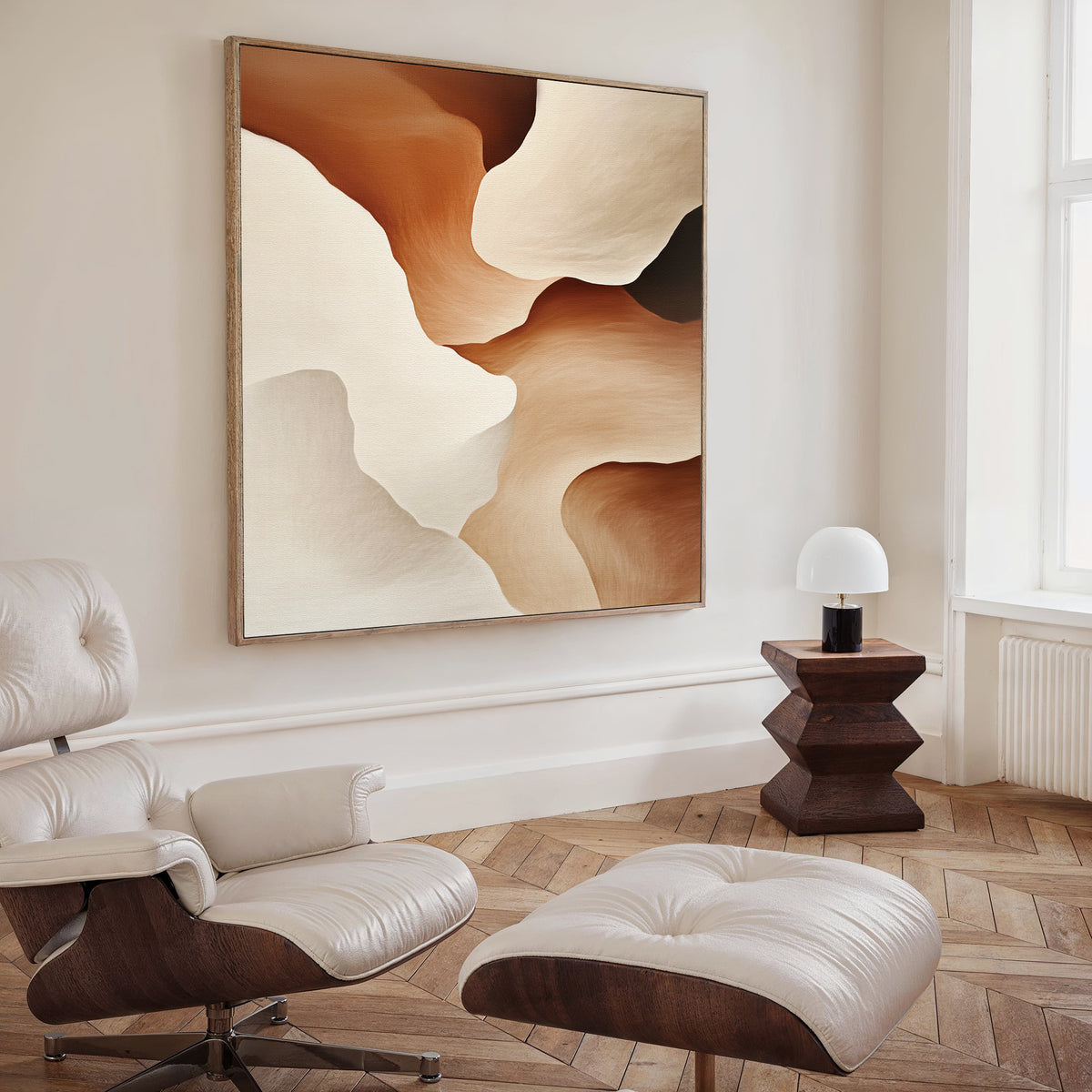 A modern living room features a white leather lounge chair and ottoman, a geometric dark wood side table, a small lamp, and the Canyon Curves Canvas Art - Square—an abstract painting in warm beige and brown tones—to complete the look.