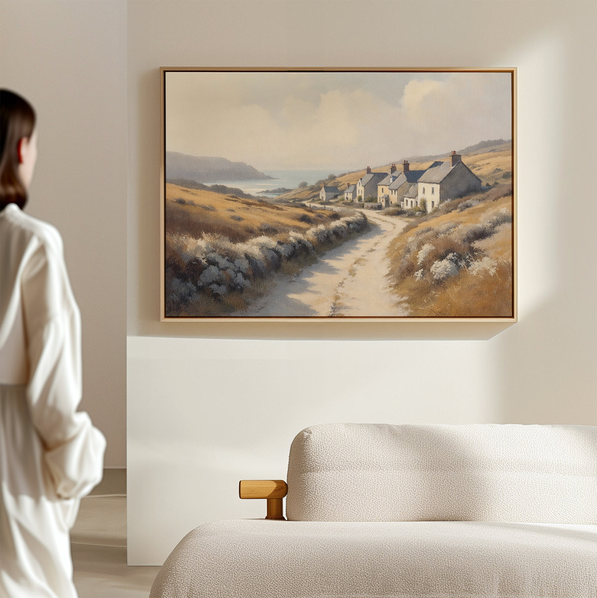 A woman stands in a minimalistic room, gazing at the Coastal Haven Canvas Art - Horizontal, a large framed print of cottages and golden fields, displayed above a beige sofa.