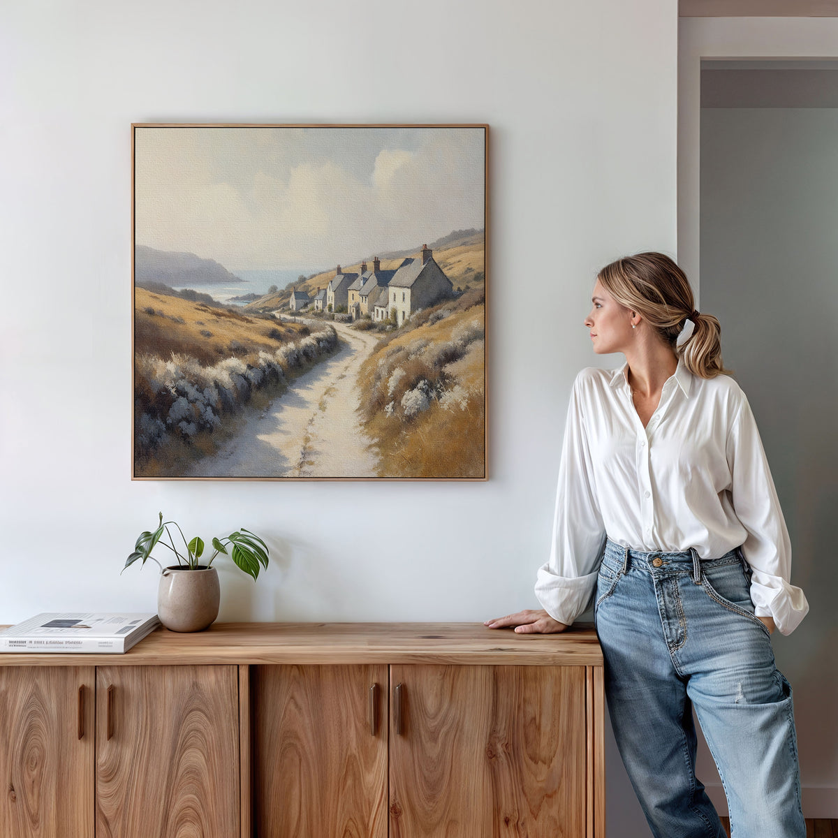 A woman in a white blouse and jeans leans against a wooden cabinet, gazing at the Coastal Haven Canvas Art - Square displayed on the white wall above her. A potted plant and books are arranged on top of the cabinet.