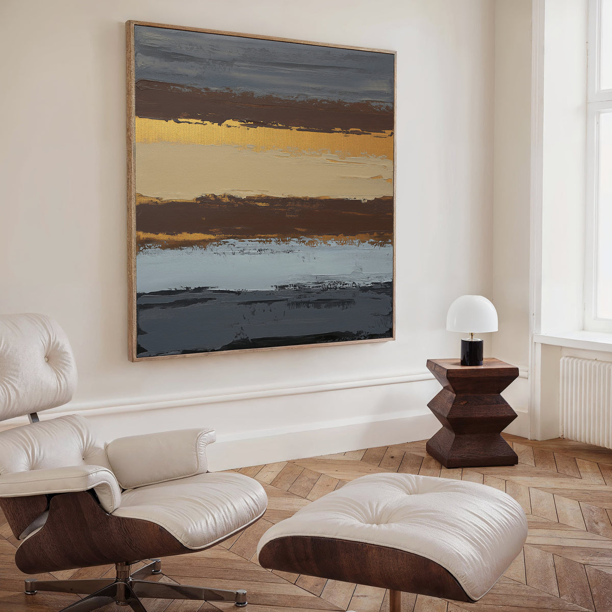 A modern living space featuring a cream Eames lounge chair and ottoman, a wood side table with a white lamp, and the Coastal Strata Canvas Art - Square showcasing horizontal earthy bands of brown, gold, cream, and blue sophistication.