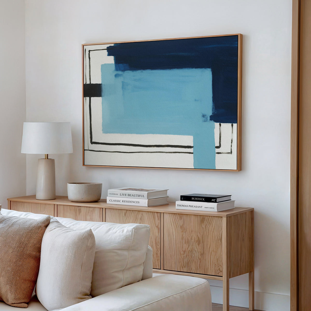 A modern living room features a wooden sideboard with books, a lamp, and a bowl. Above it hangs the Cobalt Intersection Canvas Art - Horizontal in blue and black tones. A white sofa is partly visible in the foreground.