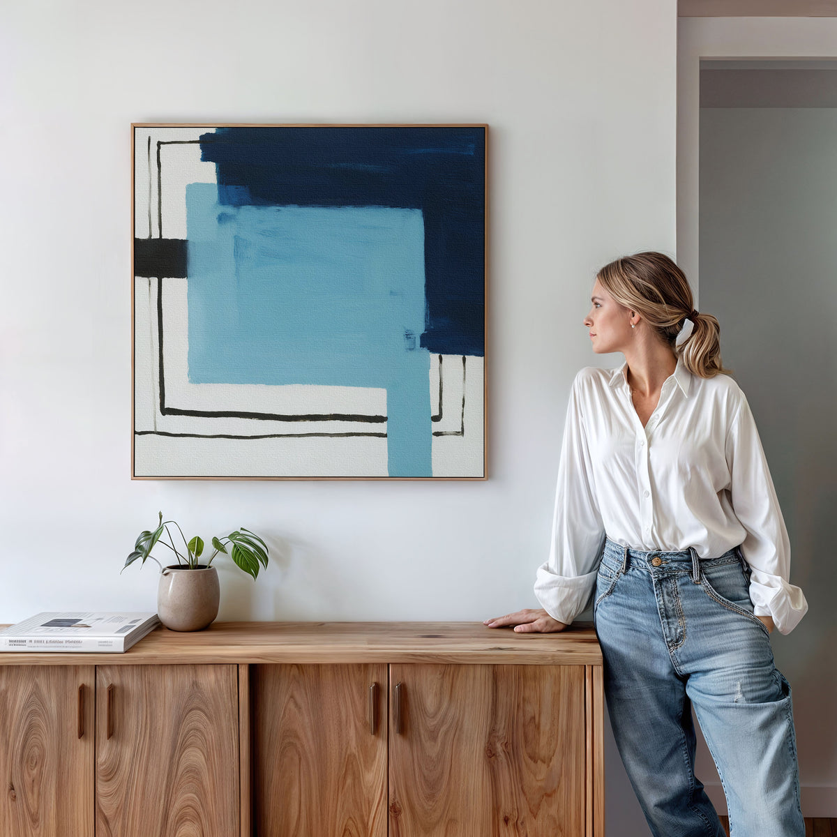 A woman in a white blouse and jeans leans against a wooden cabinet, gazing at the Cobalt Intersection Canvas Art - Square with blue and black geometric shapes on a white wall. A small plant and books rest on the cabinet.