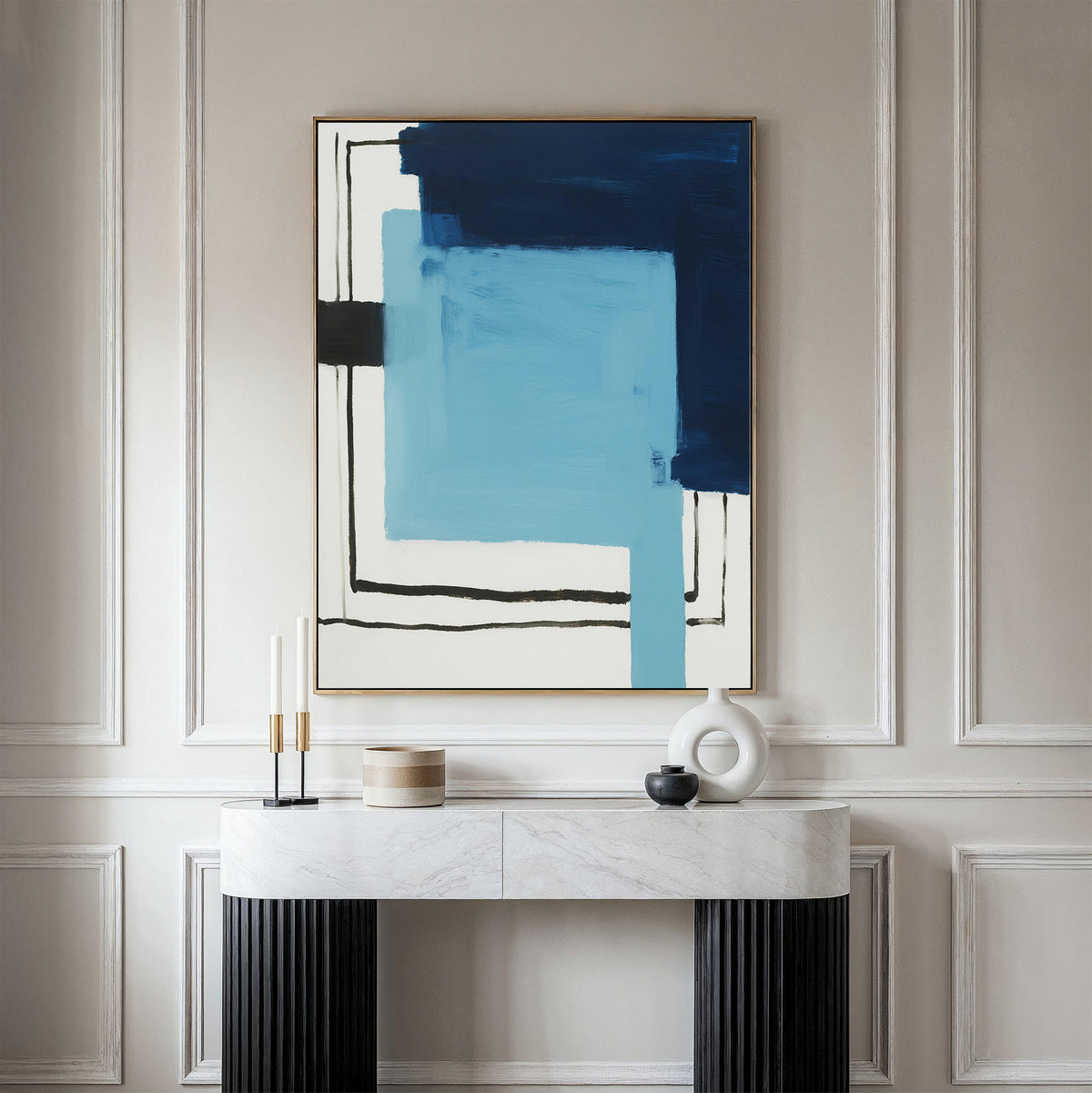 A minimalist console table with black legs and a white top displays modern decor, beneath the Cobalt Intersection Canvas Art – Vertical in blue, black, and white tones on a paneled beige wall.