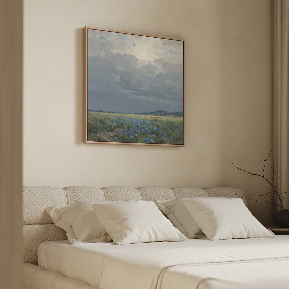 A neatly made bed with cream bedding sits beneath the Cornflower Fields Canvas Art - Square—a framed landscape print of a cloudy sky and green field with blue cornflowers—in a softly lit, minimalist bedroom.