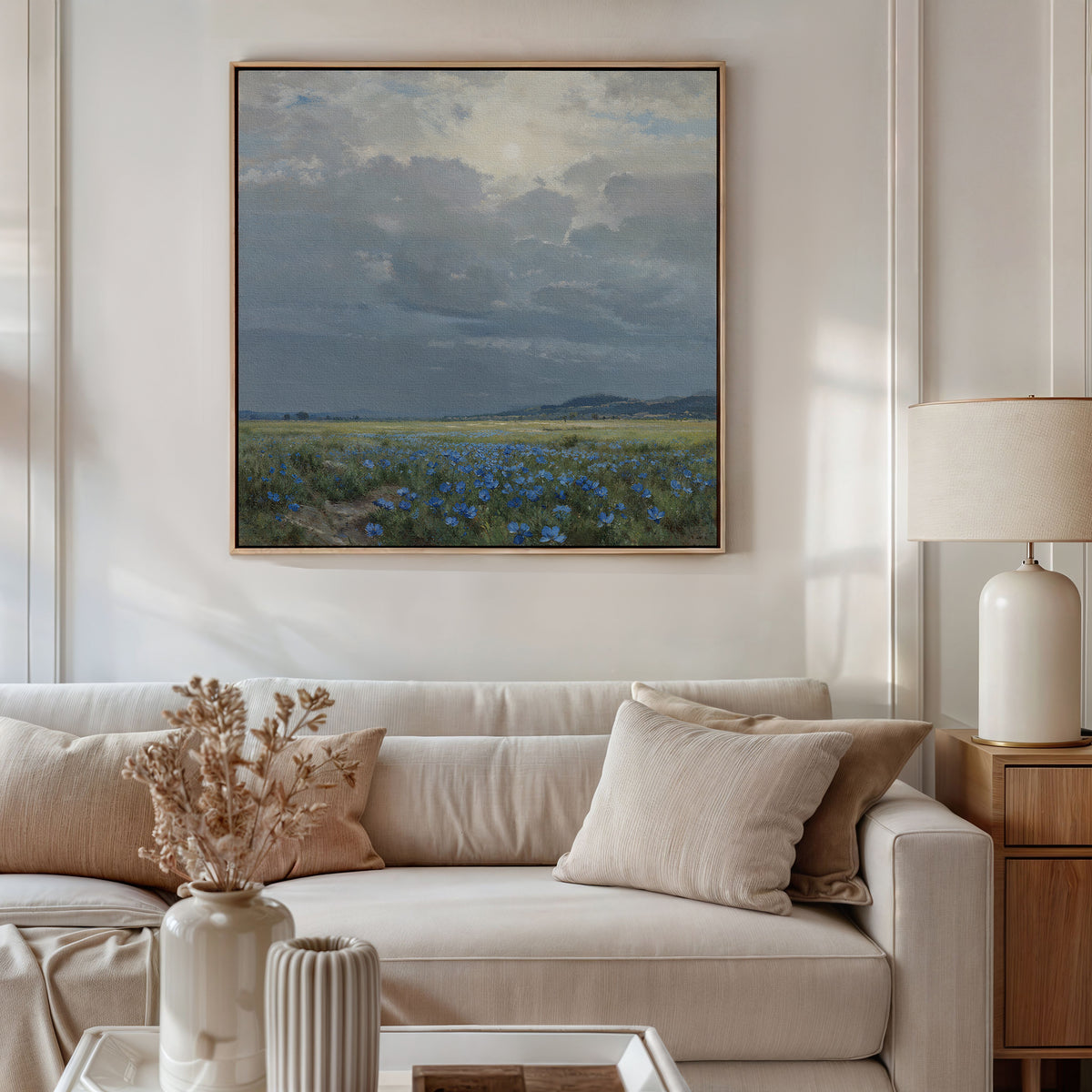 A beige sofa with neutral cushions sits against a white wall. Above it hangs the Cornflower Fields Canvas Art - Square, showing a cloudy sky over fields. A side table holds a lamp and a vase with dried flowers.
