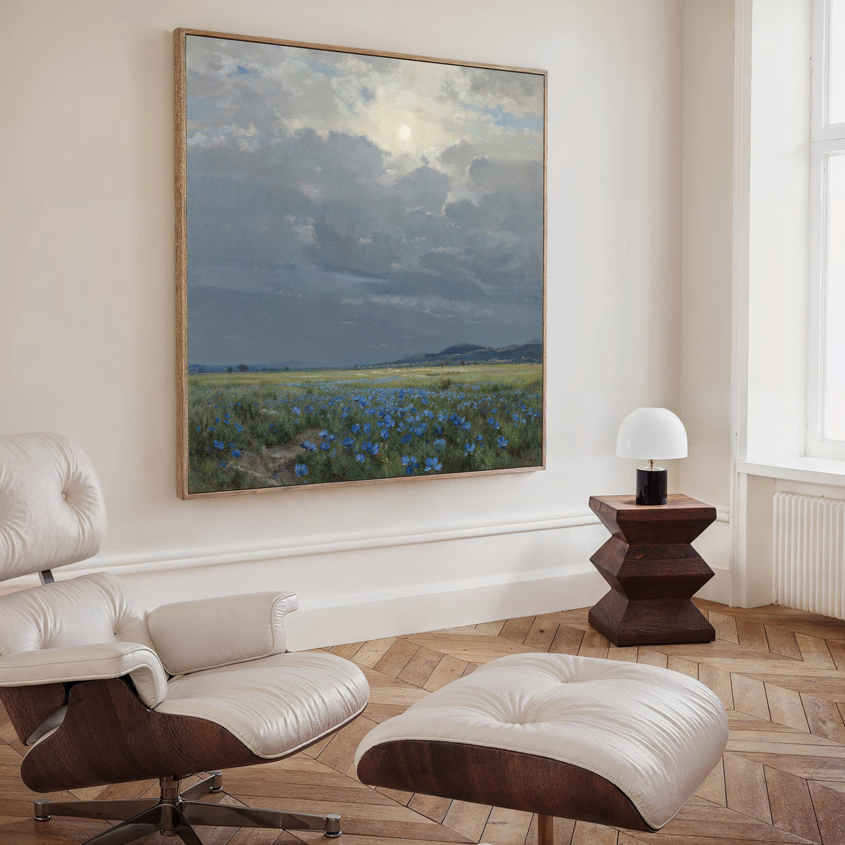 A modern living room features a white leather lounge chair with ottoman, dark wood side table and lamp, plus the Cornflower Fields Canvas Art - Square depicting a cloudy sky and fields on the wall.